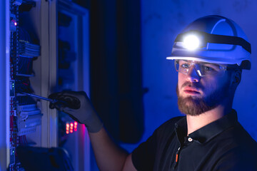 A male electrician works in a switchboard in overalls against the backdrop of emergency lighting.