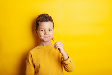 A young boy wearing a yellow sweatshirt against a bright yellow background playfully holds a red lollipop, sticking his tongue out in anticipation. The scene exudes energy and joy with vibrant tones.