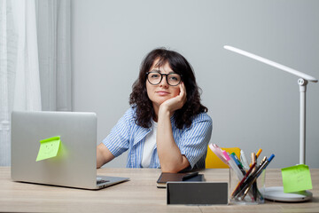 A woman sits at a desk, appearing thoughtful and slightly bored, resting her head on her hand. Wearing glasses and a striped shirt, she is surrounded by a laptop, stationery,