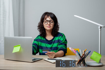 A woman wearing glasses and a green-striped shirt sits at a desk with a laptop. She holds a credit card in her hand and smiles while looking at the screen. The workspace is neat and modern