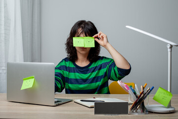 A woman in a green-striped shirt sits at a desk with her head resting on her hand. Sticky notes with drawn eyes are on her face, pretending she's awake while tired. The setting is a modern workspace.