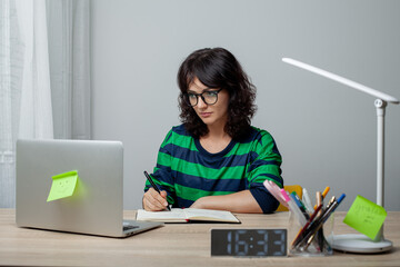 A woman in glasses and a green-striped shirt sits thoughtfully at a desk, her chin resting on her hand. A laptop, pens, and a digital clock showing 16:16 are arranged neatly on the desk