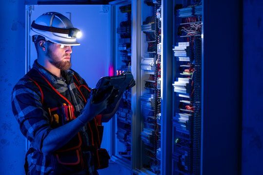 Electrician in overalls, focused on work in switchboard with fuses, using tablet.