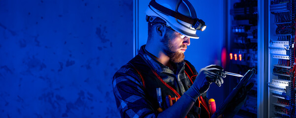 A male electrician in overalls, focused on work in switchboard with fuses, using tablet.