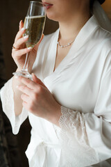 bride in a white robe standing by the window. She holds a glass of champagne and takes a sip.