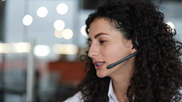 Friendly businesswoman with curly hair talks on headset in modern office. Woman enjoys pleasant conversation at workplace. Corporate communication and client service concept.