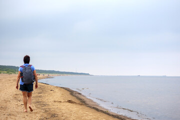 a man walking alone on a wide, sandy beach, with other people in the background and a forest in the distance.