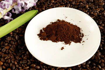 Ground coffee on a white plate on a bed of coffee beans with a purple hyacinth.