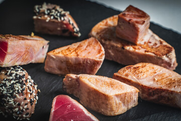 A variety of fried tuna pieces. Tuna with white and black sesame seeds and cut slices on a black mica board.