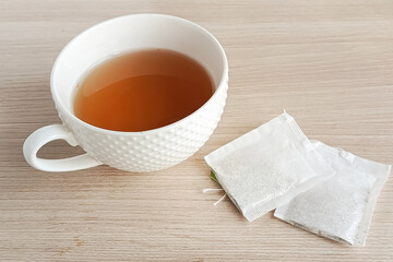 A white mug of hot tea and two tea bags on a wooden table. The benefits of black, green tea bags.