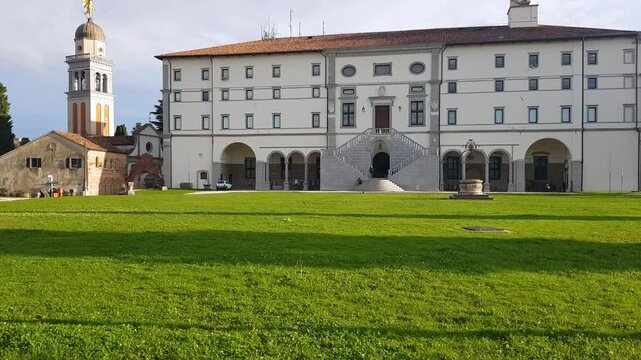 Revealing shot of Udine Castle and the Santa Maria di Castello Church, framed by the lush green grass of the square in front, on a partly cloudy autumn day