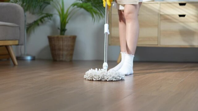 Woman wearing yellow rubber gloves and white socks is cleaning hardwood floor with a modern spinning mop in a cozy, bright, and airy apartment