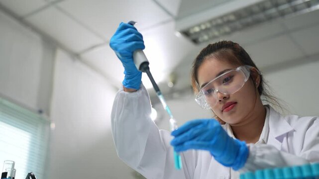Professional female scientist wearing lab coat, gloves and goggles, carefully using micropipette and analyzing green liquid in test tube, performing experiments in modern laboratory