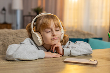 Young girl lying on table listening to an audiobook with headphones, reacting with excitement,...