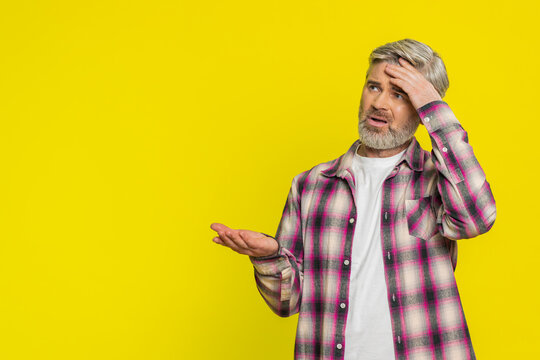 Caucasian middle-aged mature man on yellow background placing hand on forehead, heavy sigh indicating weariness. Disappointment, repeated failures creating visible irritation, comedic facepalm style