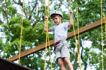 A child in a rope park in nature learns to rock climb. A boy in a helmet walks along a ropeway overcoming obstacles.