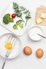 Broken egg with flour in bowl. Whisk on bowl. Butter on paper. Broccoli, tomatoes and dill