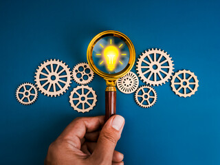Creative idea and inspiration searching tools, AI solution management workflow concept. Yellow lightbulb icon in magnifying glass lens in human hand with many gear and cogwheel, blue background.