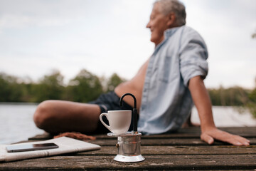 Senior man sitting on jetty at a lake with small espresso maker