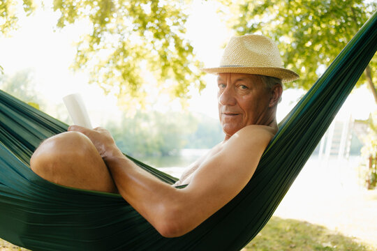 Senior man wearing straw hat relaxing in hammock at lakeshore