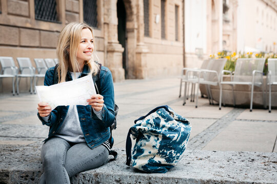 Smiling blond woman with map and baggage sitting on curb looking around