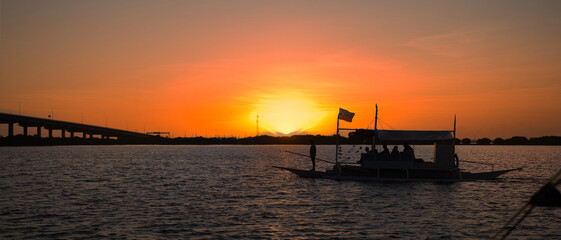 Fototapeta premium Silhouette of a traditional Philippine boat filled with passengers, floating on calm waters during a vibrant sunset, creating a serene and picturesque scene