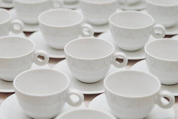 white empty porcelain cups on saucers with spoons in several rows on white tablecloth, serving tables for banquet in restaurant or cafe, pattern, background