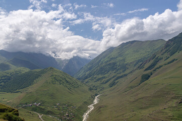 Obraz premium Panorama in the mountains with a view of the village and the mountain river. Water flows down from the peaks of the mountains, forming a river. There are white clouds over the mountains.