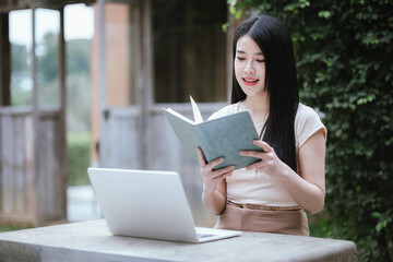A confident young Asian woman is working on her laptop in a vibrant park. E-banking and career aspirations in a modern lifestyle.