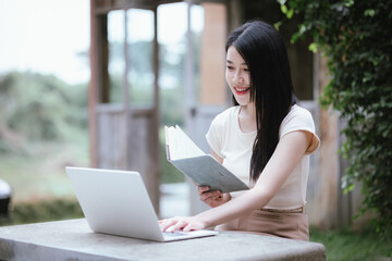 A confident young Asian woman is working on her laptop in a vibrant park. E-banking and career aspirations in a modern lifestyle.