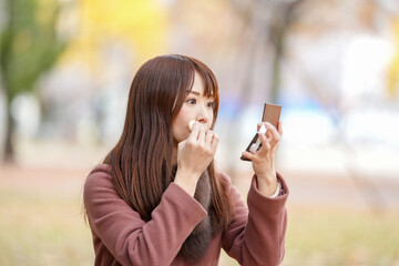 A woman with a long hair in her thirties is sitting in a beautiful autumn leaves in a park in a beautiful autumn leaves and fixing makeup