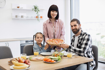 Caucasian family with parents and daughter having pizza for breakfast in bright modern kitchen. Happy child with parents sharing meal, enjoying family time and morning togetherness.