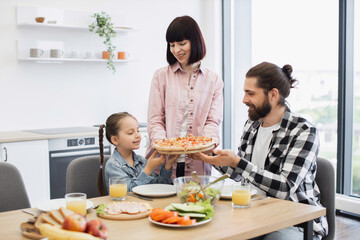 Caucasian family with young daughter enjoying pizza breakfast at home. Smiling parents and child...