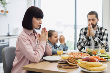Caucasian family including mother, father, and daughter praying before breakfast. Seen in modern kitchen, focused on faith and gratitude. Represents harmony and family bonding.