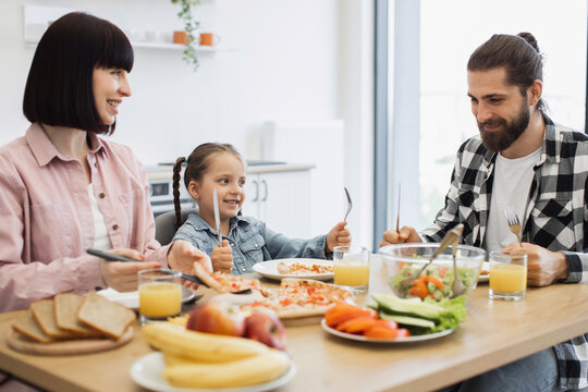 Caucasian family with young daughter enjoying breakfast together. Parents and child smiling, eating pizza and fresh salad. Bright morning scene with healthy fruits and orange juice.