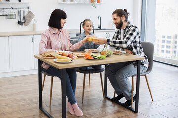 Caucasian family with father, mother, and daughter clinking glasses during breakfast. Happy mealtime in bright kitchen setting with fresh food and orange juice. Family bonding and morning routine.
