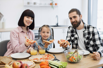Caucasian family with parents and daughter having breakfast with pizza. Family smiles while dining together in bright kitchen, showing happiness and togetherness.