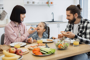 Caucasian mother and young daughter sitting together enjoying pizza for breakfast at home. Warm family moment with orange juice and smiling faces.