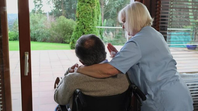 Nurse comforting elderly man in wheelchair looking at garden