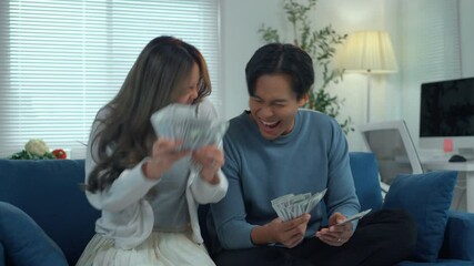 Young asian couple counting dollar bills while sitting on home sofa, expressing excitement, throwing cash, celebrating financial achievement