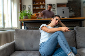 Smiling woman enjoys morning television from cozy sofa while partner handles breakfast preparation...