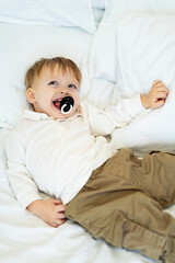 Child enjoying playtime on bed. A joyful young boy with a pacifier smiles while lying on a cozy...