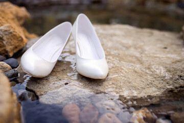 Elegant white shoes resting by water. A pair of elegant white shoes rests on a rock near clear water, surrounded by stones and nature.