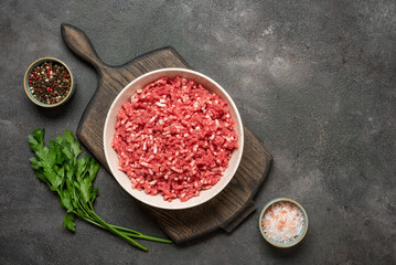 Raw mince beef, ground meat in bowl with spices on wooden cutting board, dark grunge background,...