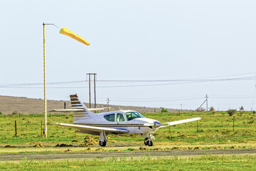 White and brown four-seat single engine light aircraft parked next to windsock at a small airstrip...