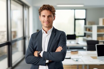 Fototapeta premium Portrait of a smiling young businessman standing with arms crossed in a modern office, exuding confidence and professionalism