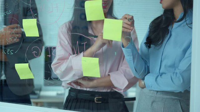 Strategic brainstorming session with businesswomen collaborating, writing and drawing ideas on glass wall using sticky notes during dynamic team planning meeting