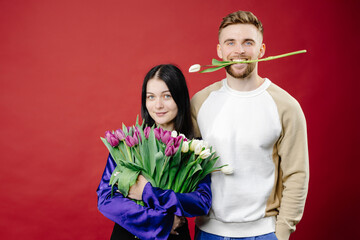 Shocked lady and positive man holding white fresh tulips, looking at camera isolated on blue...