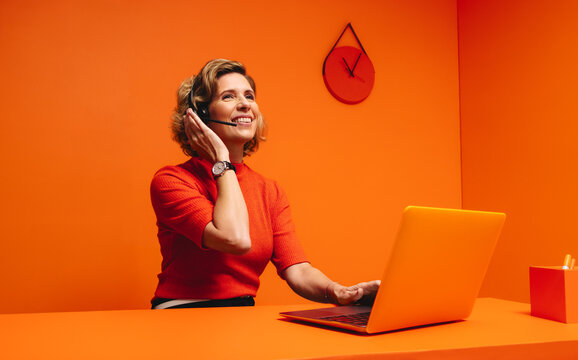 Smiling mature woman with headset in monochromatic orange workspace, emphasizing creativity and modernity