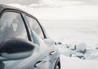 Obraz premium Traveling in winter - part of the car on the bank of huge frozen lake. Left side of silver SUV on snow on sunny afternoon. 